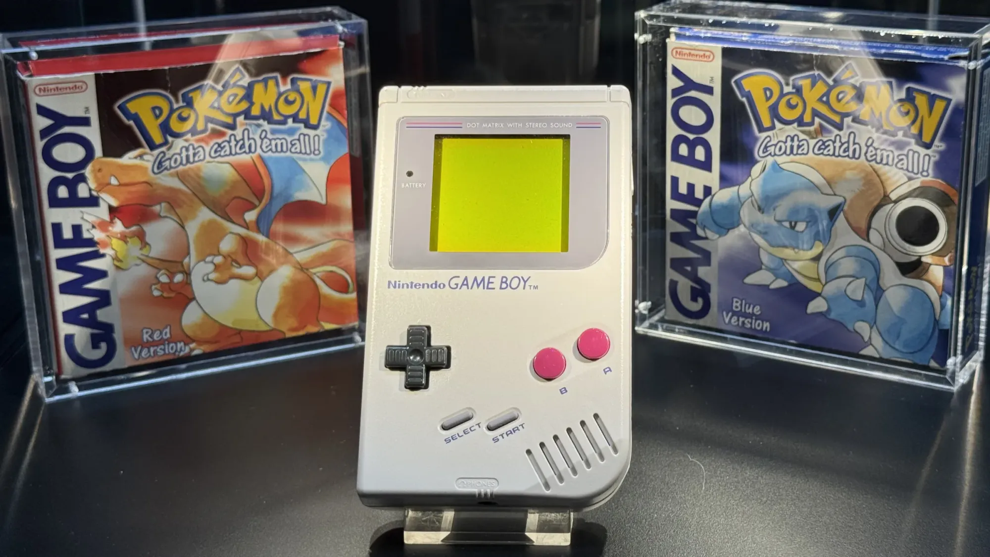 Pokémon Red and Blue boxes flanking an original Nintendo Game Boy inside a glass case.